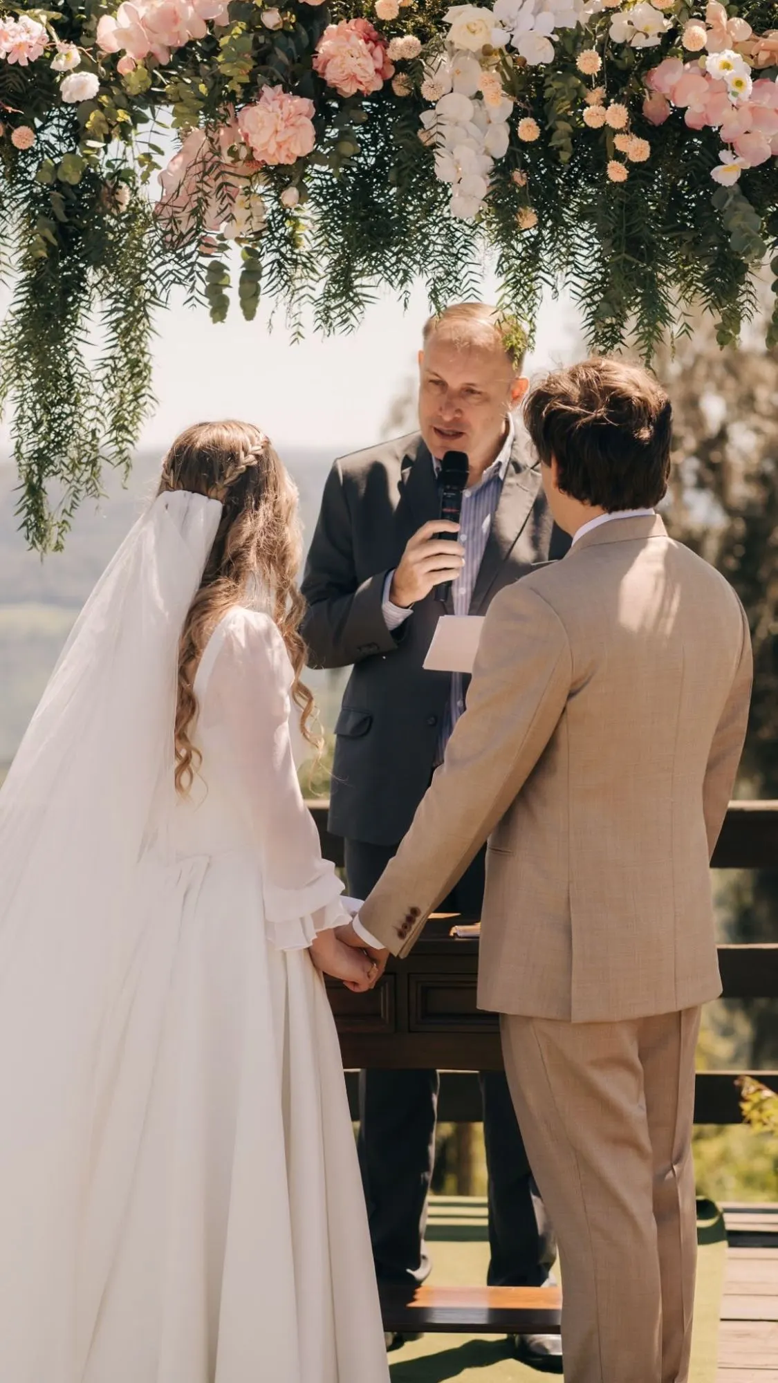 Noivos de mãos dadas diante de Ricardo, sob arco de flores rosadas