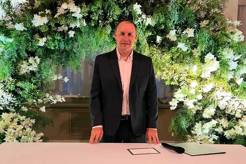 Retrato de Ricardo Backes diante de um painel de folhagens e flores brancas